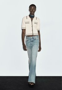 Top in maglina color crema con bordi neri e bottoni dorati, abbinato a jeans svasati azzurri. Modello stilizzato con una sciarpa blu e scarpe aperte.
