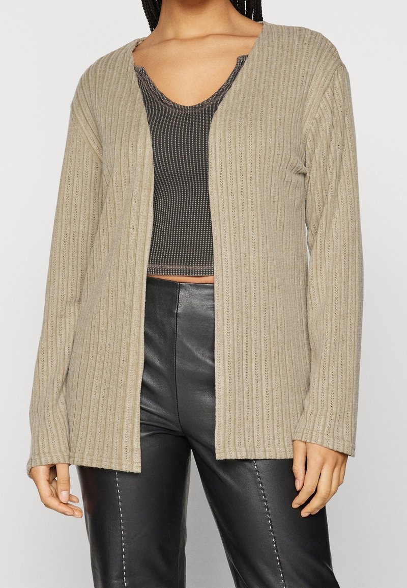 Beige ribbed cardigan with an open front, long sleeves, and textured fabric, layered over a fitted black top and paired with black pants.