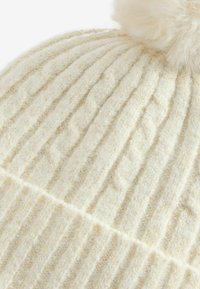 Cremefarbene gestrickte Mütze mit vertikaler Rippstruktur, ausgestattet mit einem weichen Kunstfell-Pompon oben und dezenten goldenen Akzenten.