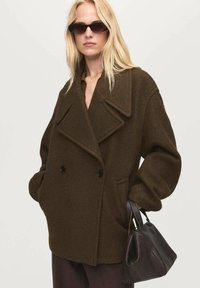 Brun uldblandet dobbeltradet frakke med store kraver og oversized ærmer, parret med en lille mørk lædertaske. Synlige knapper i gunmetal.