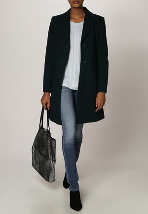 Donkergroene wollen jas met een reverskraag en dubbele knoopsluiting, gedragen over een lichtblauwe kabelgebreide trui, gecombineerd met een blauwe spijkerbroek en een zilveren tas.