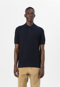 Polo de culoare albastră închis, cu mânecă scurtă, fabricat dintr-un material texturat, cu guler cu trei nasturi și manșete ribbate, purtat cu pantaloni de culoare deschisă.