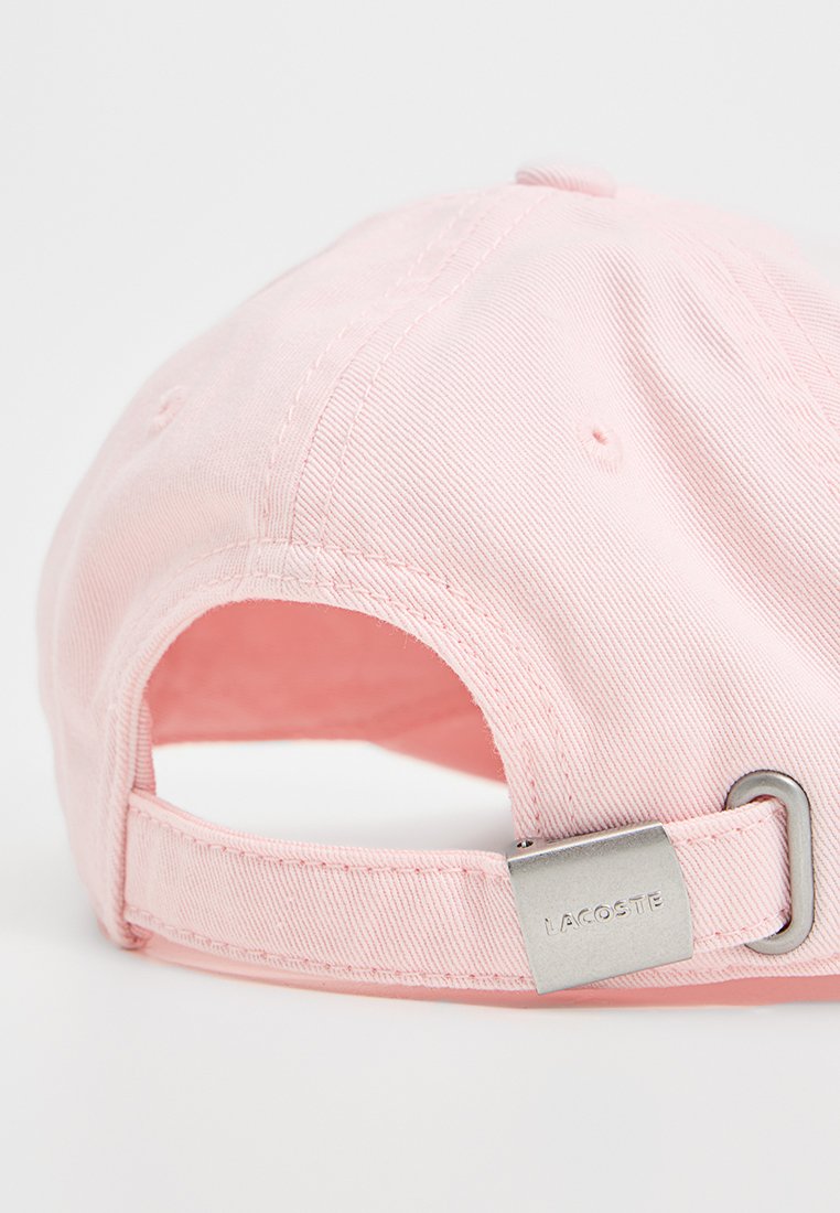 Gorra de algodón rosa con una correa ajustable. La tela tiene una textura acanalada. El herraje de metal plateado presenta el logo "LACOSTE."