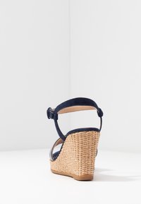 Námořnické modré semišové sandály na klínu s tkaným slámovým podpatkem, kotníkovým řemínkem a otevřeným zadním designem. Hladká textura a přírodní akcenty vyznačují tuto obuv.