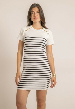 Femme portant une robe blanche à manches courtes avec des rayures horizontales noires et des boutons dorés sur les épaules, debout devant un fond uni.