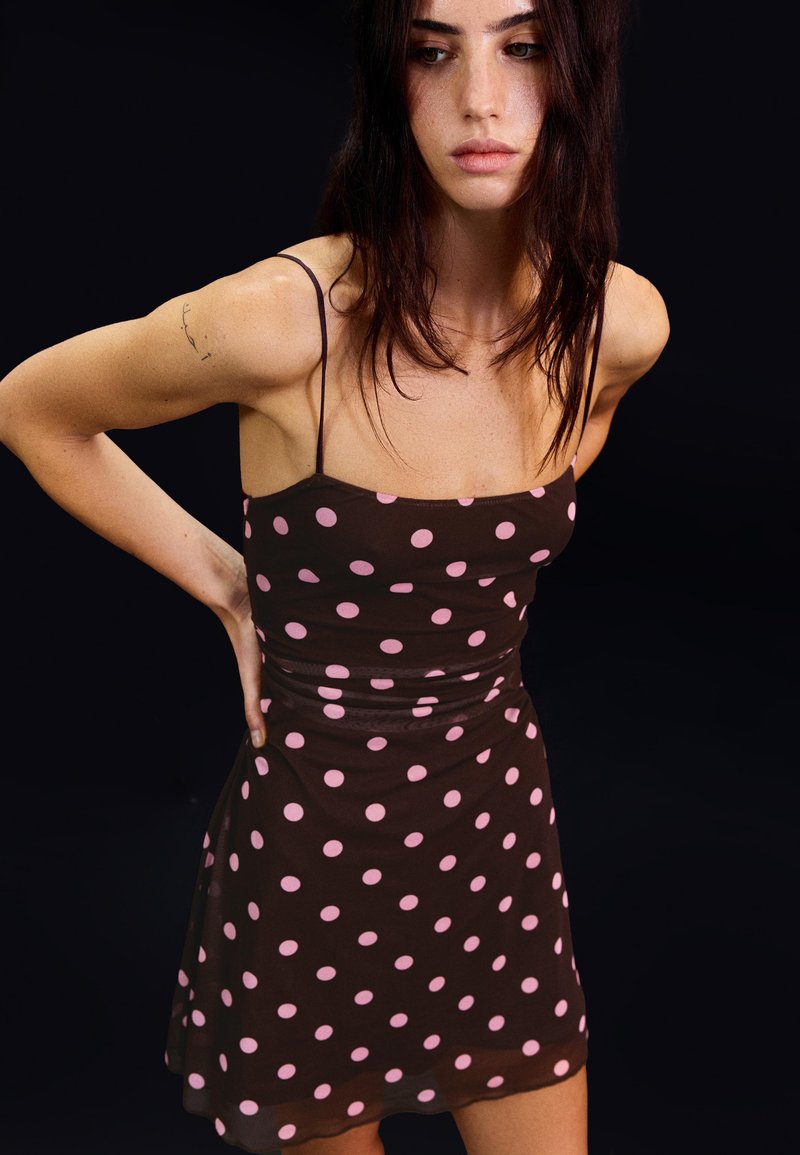 Kobieta o ciemnych włosach, ubrana w brązową sukienkę bez rękawów w różowe kropki, stojąca na czarnym tle, patrząca w dół.