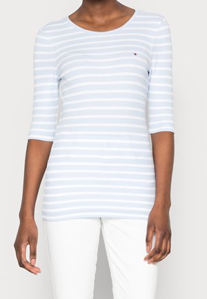 Kobieta ubrana w jasnoniebieską i białą koszulę w paski z rękawami do łokcia i małym logo na piersi, w połączeniu z białymi spodniami.