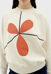 Sweatshirt crème avec un grand motif floral abstrait rouge, des détails noirs et des poignets côtelés. Fabriqué en tissu doux et texturé.