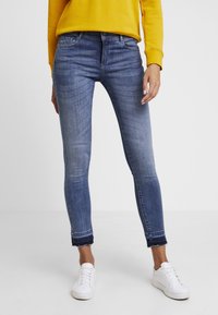 Henkilö, jolla on yllään haalistuneet siniset nilkkahousut, valkoiset tennarit ja keltainen pitkähihainen paita, seisoo valkoista taustaa vasten.