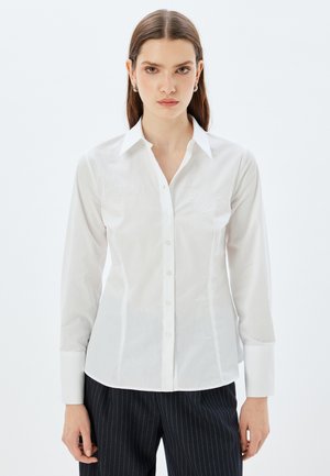 Camisa blanca de manga larga, con botones, fabricada en una tela suave, que presenta un collar puntiagudo y un sutil bordado en el pecho.