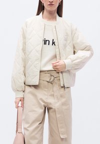 Μπεζ bomber τζάκετ με καπιτονέ σχέδιο και ριμπ μανσέτες, συνδυασμένο με κρεμ πουλόβερ και μπεζ παντελόνι, κρατώντας μια απαλή ροζ τσάντα.