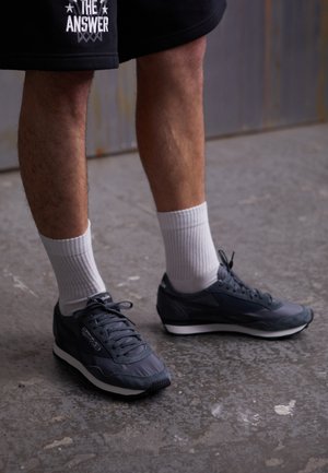 Personne portant des baskets Reebok noires, des chaussettes blanches montantes et un short noir avec le texte "THE ANSWER", debout sur un sol en béton gris.