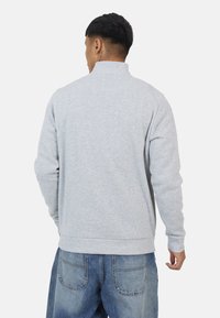 Felpa grigia con colletto alto, maniche lunghe e polsini a costine. Presenta una texture morbida e una vestibilità rilassata, indossata con jeans di denim blu.