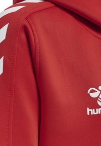 Rode sportjack met witte chevronstrepen op de schouders en een gedeeltelijk wit "hummel" logo zichtbaar op de borst.