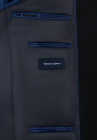 Binnenkant van een jas met een gladde zwarte voering en donkere blauwe accenten, meerdere zakken, en een label met de tekst "THOMAS GOODWIN".