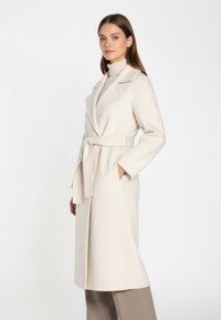 Cappotto lungo con cintura in misto lana bianco sporco, dotato di colletto a scialle, due tasche e una finitura testurizzata. Indossato sopra un leggero dolcevita.