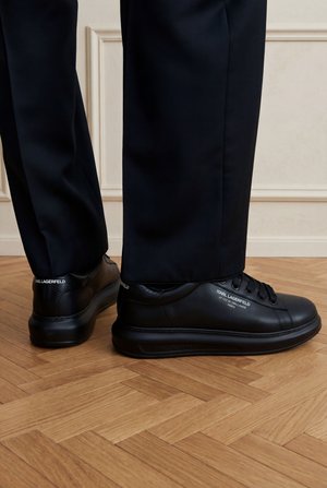 Osoba nosiaca čierne tenisky Karl Lagerfeld s čiernymi nohavicami, stojaca na drevenej parketovej podlahe proti bielej doskovanej stene.