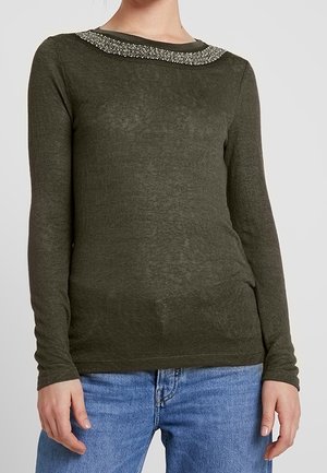 Personne portant un haut vert foncé à manches longues avec un ornement argenté texturé le long de l'encolure et un jean en denim bleu.