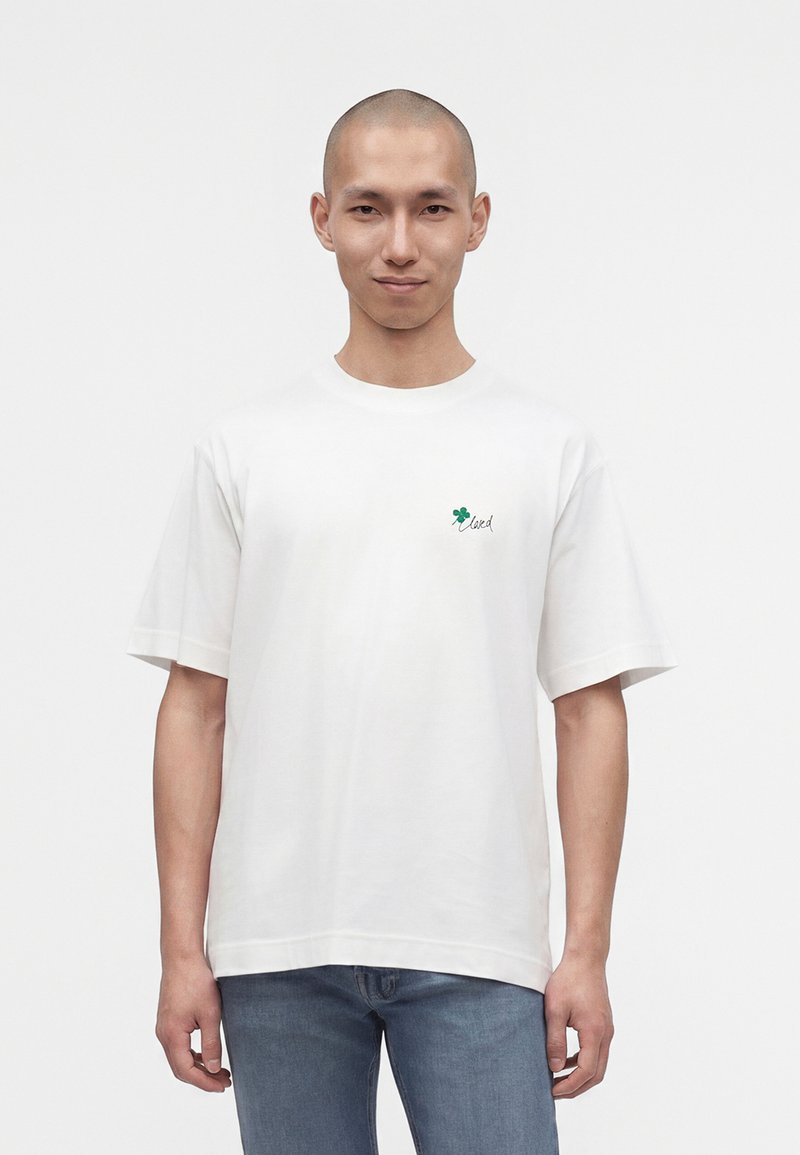 Jeune homme au crâne rasé porte un T-shirt blanc avec un petit trèfle vert et le mot "loved" sur la poitrine, associé à un jean bleu, debout sur fond blanc.