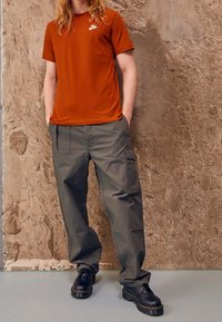 Oranžové tričko s krátkým rukávem a bílým logem; šedé cargo kalhoty s bočními kapsami; černé šněrovací boty s širokými podešvemi.