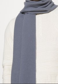 Écharpe en laine grise avec une texture douce, drapée autour du cou, contrastant avec un gros pull en tricot blanc.