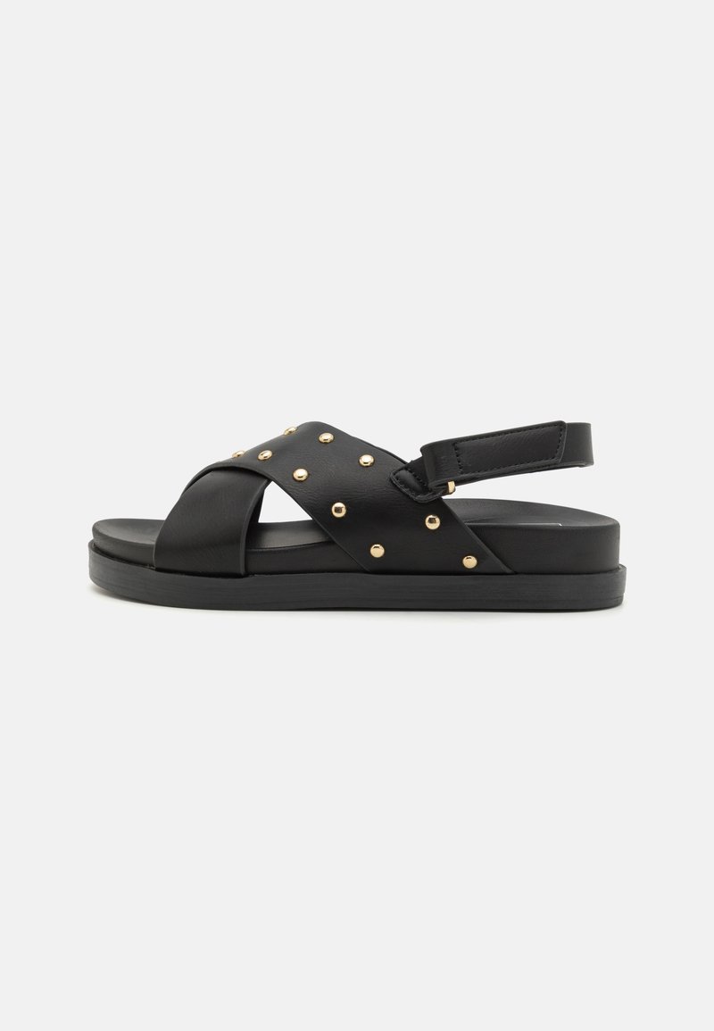 Sandale en cuir noir avec un design de bride croisée, ornée d'accents en clous dorés et d'une sangle de cheville réglable. Semelle en caoutchouc plate.