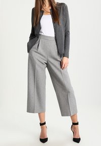 Grijze wollen wijde broek gecombineerd met een grijze cardigan en een witte top met spaghettibandjes, met een relaxte pasvorm en een gedefinieerde taille. Zwarte hakken met enkelband.