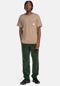 Tan cotton t-shirt with a pocket detail, paired with dark green corduroy pants. Black athletic shoes complete the outfit.