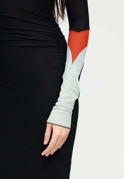 Κοντινό πλάνο ενός ατόμου που φοράει μαύρο φόρεμα με ρυτιδωτό ύφασμα και μακριά μανίκια με μπλοκάρισμα χρωμάτων σε κόκκινο και ανοιχτό μπλε.