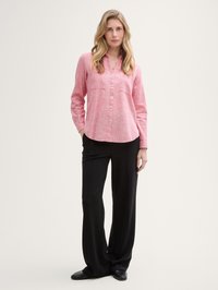 Camicia button-up rosa chiaro con scollo a V e maniche lunghe, abbinata a pantaloni neri a gamba larga e scarpe nere.