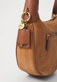 Bolso de ante marrón con parte superior de cuero liso, herrajes dorados, bolsillo con cremallera y una pequeña etiqueta de cuero con un logo.