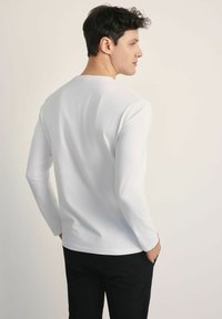 Biała bluzka z długim rękawem, wykonana z gładkiego materiału, z okrągłym dekoltem i dopasowanym krojem. Noszona z czarnymi spodniami, pokazana z tyłu.