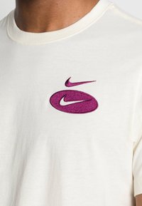Kremowa bawełniana koszulka z okrągłym dekoltem, z dużym haftowanym fioletowym logo Nike z przodu. Krótkie rękawy, miękka faktura.