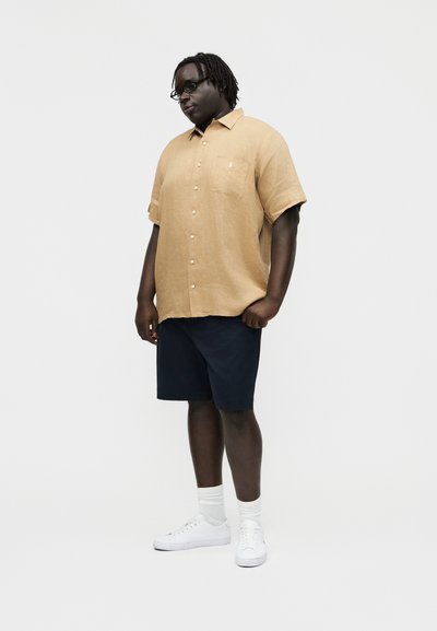 Mees prillidega, kandes lühikeste varrukatega beeži värvi nööbitavat särki, mereväe siniseid lühikesi pükse, valgeid sokke ja valgeid tossusid, seisab ühevärvilise tausta ees.