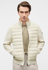 Ανοιχτόχρωμο μπουφάν με λεπτομέρειες καπιτονέ στο μπροστινό μέρος, πλεκτά μανίκια και ψηλό γιακά. Κλείνει με φερμουάρ, διαθέτει ριμπ μανσέτες και λογότυπο στο αριστερό μανίκι.