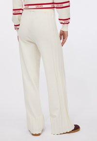 Pantaloni larghi color crema con una texture a coste, caratterizzati da strisce laterali e fondo svasato. Dettaglio logo dorato sulla cintura.