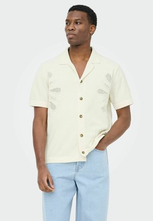 Férfi, aki rövid ujjú krémszínű gombos ingben, leveles kivágásmintákkal, világoskék farmert visel, kezét zsebébe téve áll.