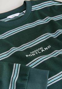 Tamsiai žalia ilgomis rankovėmis marškinėliai su horizontaliomis šviesiai mėlynomis ir baltomis juostelėmis, kuriuose balta siuvinėta užrašas "OREGON PORTLAND". Minkšta medvilnės medžiaga.