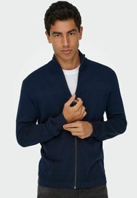 Cardigan zippé marine présentant des motifs de tricot texturés, un col et des poignets côtelés, et une fermeture éclair lisse. Porté par-dessus un t-shirt blanc.