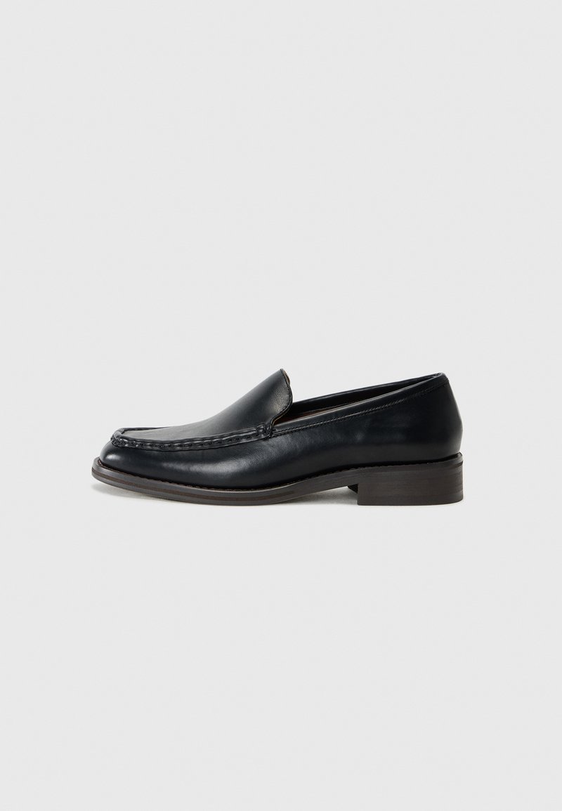 Mocasín de cuero negro para hombre con puntera mocasinada cosida, tacón de madera bajo y diseño deslizante sobre un fondo claro y uniforme.