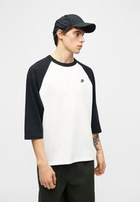 Νέος άνδρας που φοράει μαύρο καπέλο και λευκό μπλουζάκι μπέιζμπολ Nike με μαύρα μανίκια, στέκεται πλάγια μπροστά σε απλό ανοιχτό φόντο.