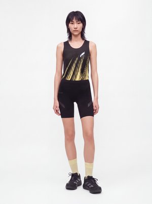 Jeune personne debout, vêtue d'un haut sans manches noir avec un motif jaune, un short de cycliste noir, des chaussettes jaune clair et des baskets noires sur fond blanc.