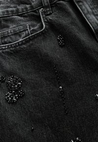 Mustikka-denim-kangas, jossa on teksturoitu pinta ja mustathelmikoristeiset kukkaelementit sekä vähäistä hohtoa. Taskut ja ompelusyys ovat näkyvissä.