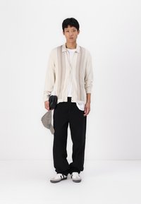Light beige cardigan with vertical ribbing detail, white t-shirt underneath, black loose-fitting pants, and gray sneakers, held cap.