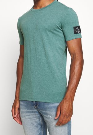 Kurzärmeliges T-Shirt mit Rundhalsausschnitt aus meliertem grünem Stoff, mit einem schwarzen Calvin Klein Logo-Patch am linken Ärmel. Glatte Textur.