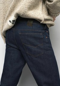 Jean en denim bleu foncé avec coutures orange, présentant une étiquette en cuir sur la ceinture et deux poches arrière avec des coutures en biais.