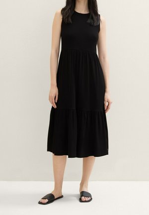 Sort ærmeløs kjole med en tætsiddende overdel og lagdelt nederdel. Fremstillet i blødt materiale, med rundet udskæring og flydende silhuet.