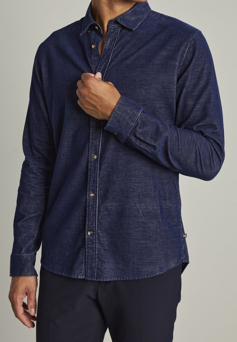 Homme portant une chemise en jean bleu foncé à manches longues en train de boutonner le bouton supérieur, montré du cou aux hanches sur un fond neutre.