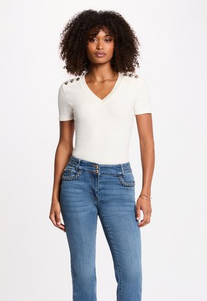 Kvinde med krøllet hår iført en cremet V-hals kortærmet top med skulderknapper og blå højtaljede jeans med flettede lommer.