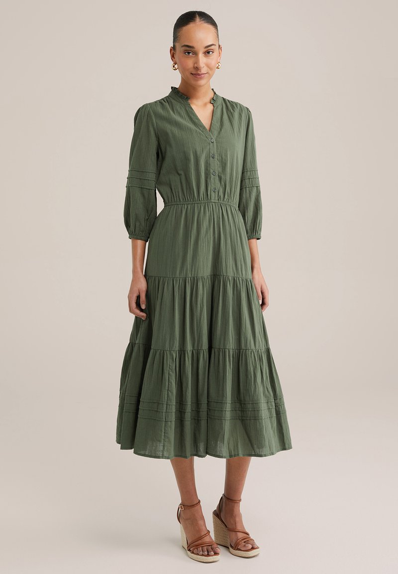 Abito lungo verde in tessuto leggero, con chiusura a bottoni sul davanti, design a strati, maniche a tre quarti e vita elasticizzata. Indossato con sandali con tacco a cuneo.
